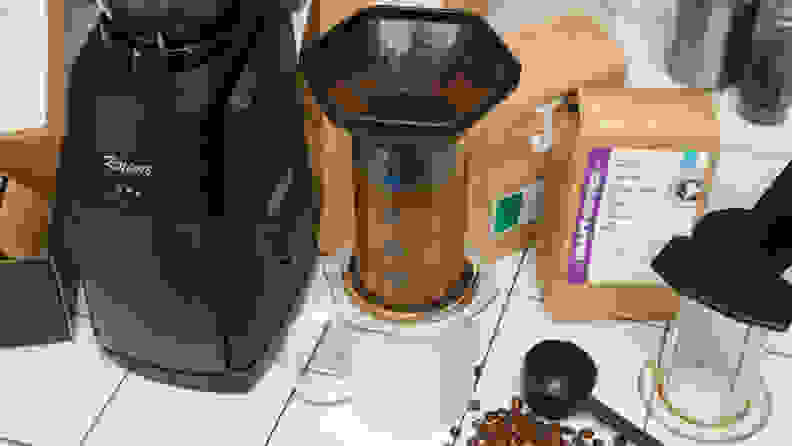 Assorted coffee gadgets arranged on a white surface, including a coffee mug, an Aeropress coffee maker, a coffee bean grinder, a measuring spoon, and coffee bean bags.