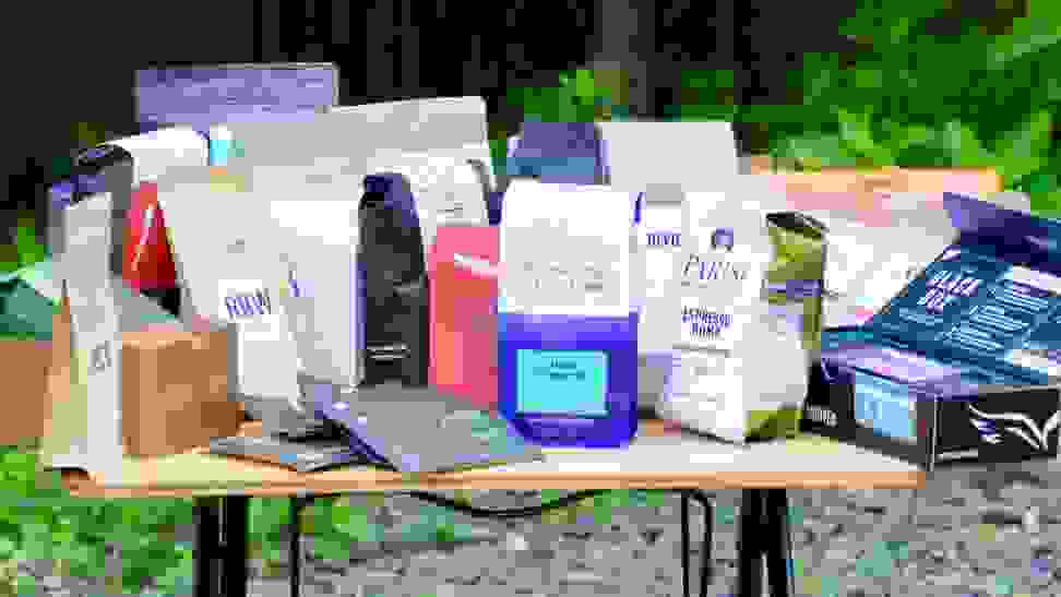 Over a dozen different bags of coffee beans arranged on a wood table outdoors.