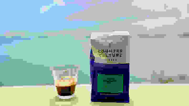 A glass espresso cup filled with a shot of espresso sits next to a bag of coffee beans on a table outside.
