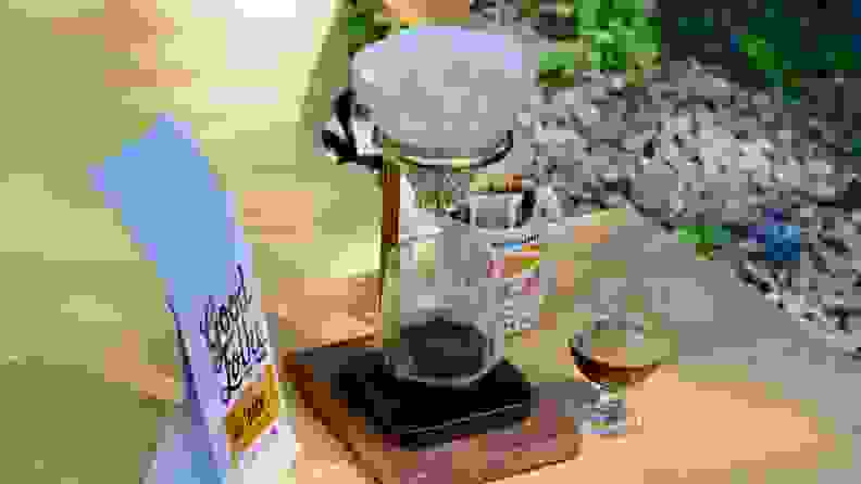 A pour-over coffee maker sits on a wooden table outside, accompanied by a bag of coffee beans and a cup filled with freshly made espresso.