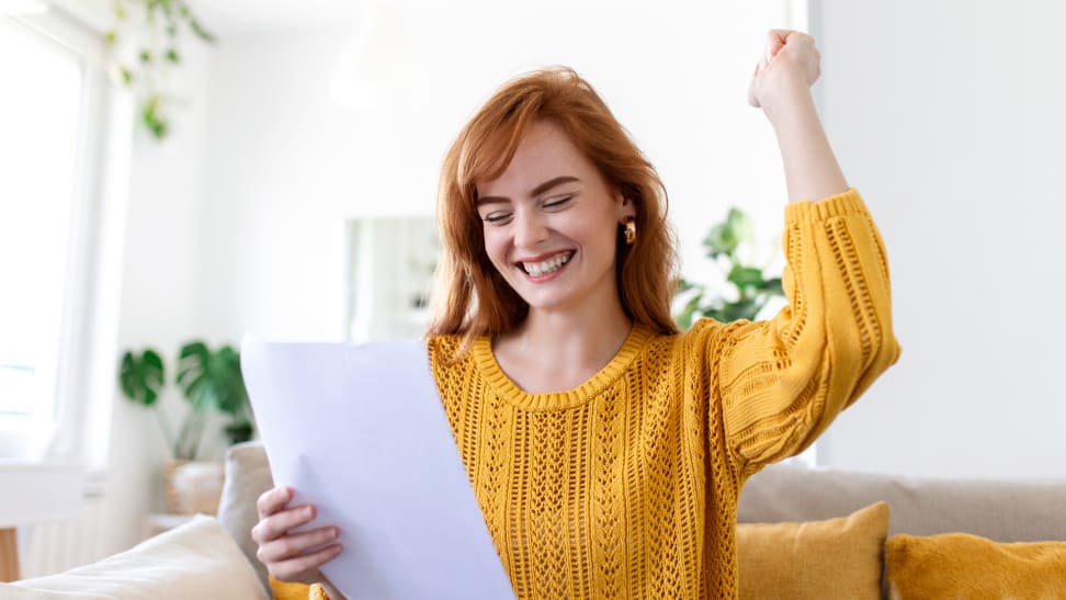 A woman in a yellow sweater smiles and raises her fist while reading a document, celebrating good news.