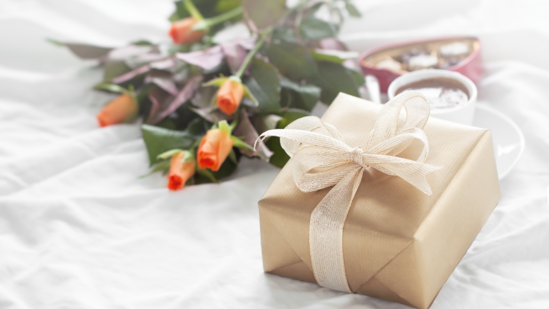 A beautifully wrapped gold gift box with a ribbon, accompanied by roses and chocolates.