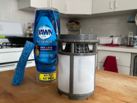 A dishwasher filter next to a bottle of dish soap and a sponge.