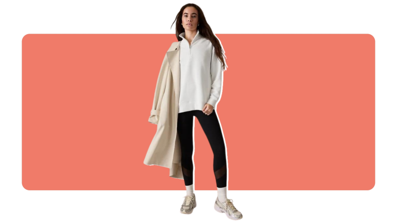 A model wearing black Salutation Stash high-rise leggings, a beige pullover and a stylish jacket resting on their shoulder with a matching pair of sneakers.