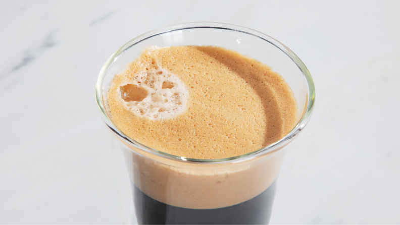 Foamy coffee in clear glass coffee mug on top of marble countertop.