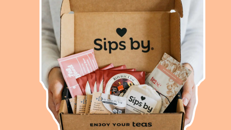 Person holding A Sips By tea gifts box featuring an assortment of teas and tea-related products with both hands.