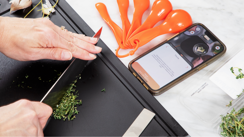 A person cutting some ingredients on a cutting board while looking at tutorial videos on a smartphone.